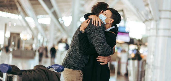 Ehemann und Ehefrau umarmen sich am Flughafen. Eine Frau mit Gesichtsmaske begrüßt ihren Mann nach der Ankunft aus dem Ausland nach der Pandemie. - JLPSF06060