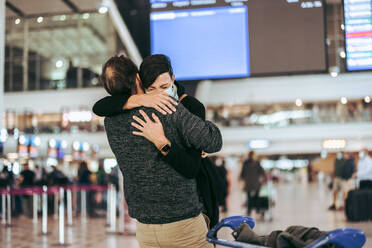 Excited woman meeting husband during pandemic at airport arrival gate. Man giving warm hug to his wife on arrival at airport in pandemic. - JLPSF06061