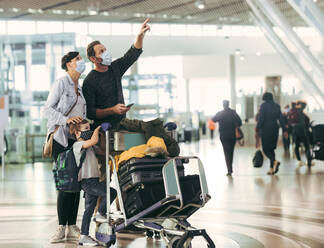 Mann zeigt auf Ankunfts-Abflugtafel und spricht mit Frau am Flughafen. Familie mit Gepäckwagen wartet auf ihren Flug am Flughafen. - JLPSF06077