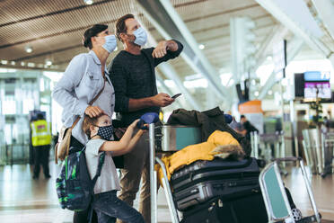 Familie mit Gesichtsmasken, die an Bord des Flughafens auf ihre Flugzeiten schaut. Tourist, der mit Frau und Kind am Flughafen wartet, mit Gepäck auf einem Trolley während einer Pandemie. - JLPSF06078