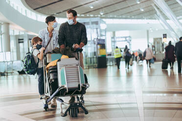 Eine dreiköpfige Familie, die mit einem Gepäckwagen im Flughafen spazieren geht, während der Pandemie. Ein Paar, das mit einem Kind im Flughafen spazieren geht, während der Pandemie. - JLPSF06083