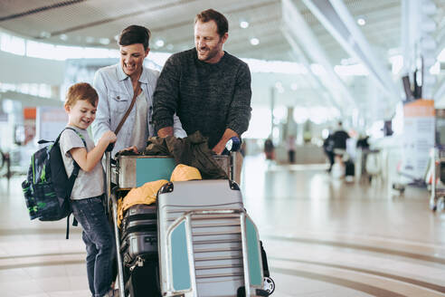 Husband and wife looking at child standing on luggage trolley at airport. Couple looking at their son at airport with luggage trolley. - JLPSF06086