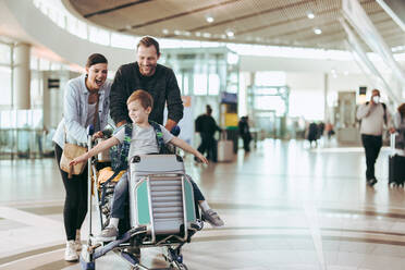 Ein Ehepaar schiebt den Gepäckwagen mit seinem Sohn am Flughafen. Ein Kind genießt es, auf dem Gepäckwagen zu sitzen, während die Eltern ihn am Flughafen schieben. - JLPSF06087