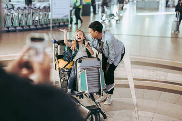 Mutter mit Sohn auf Gepäckwagen sitzend, während Vater Fotos macht. Vater fotografiert Frau und Sohn am Flughafen. - JLPSF06089