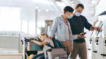 Ein Ehepaar nutzt den Selbstbedienungs-Check-in-Automaten, während ihr Kind auf dem Gepäckwagen sitzt. Eine Touristenfamilie beim Selbst-Check-in am Flughafen während der Pandemie. - JLPSF06105