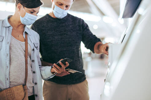 Ein Ehepaar gibt seine Daten in den Selbstbedienungs-Check-in-Automaten am Flughafen ein. Eine Touristenfamilie mit Gesichtsmasken beim Selbst-Check-in am Flughafen. - JLPSF06106