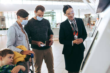Flughafenpersonal mit Ehepaar und ihrem Sohn am Selbstbedienungs-Check-in-Automaten. Das Bodenpersonal hilft einer Familie von Reisenden am Flughafen während der Pandemie. - JLPSF06108