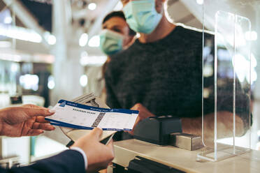 Flughafenangestellte am Check-in-Schalter mit Bordkarte und Reisepass eines Reisenden. Mitarbeiter der Fluggesellschaft helfen einem maskierten Mann während einer Pandemie am Check-in-Schalter. - JLPSF06119