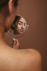 Woman with vitiligo looking at a mirror against brown background. Rear view of young woman feeling confident about her skin condition. - JLPSF06145