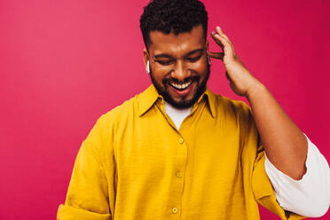 Music and happiness. Young man smiling cheerfully while while streaming his favourite music using wireless earphones in a studio. Carefree young man standing alone against a pink background. - JLPSF06226