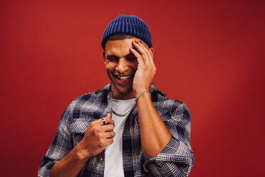 Young man with a beanie standing in a studio. Happy young man smiling cheerfully while standing against a red background. Fashionable young man wearing nail polish and eye shadow. - JLPSF06234