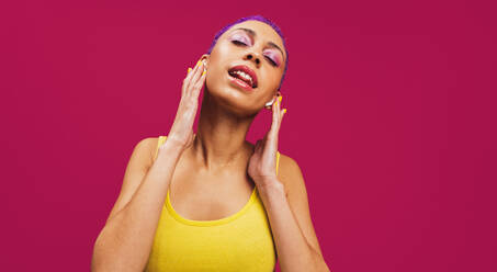 Glückliche junge Frau, die ihre drahtlosen Kopfhörer berührt, während sie ihren Lieblingssong hört. Frau mit lila Haaren steht mit geschlossenen Augen vor einem rosa Hintergrund, - JLPSF06257