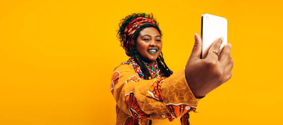 Modische junge Frau, die ein Selfie macht, während sie traditionelle afrikanische Kleidung trägt. Glückliche junge Frau, die fröhlich lächelt, während sie vor einem gelben Hintergrund steht. - JLPSF06281