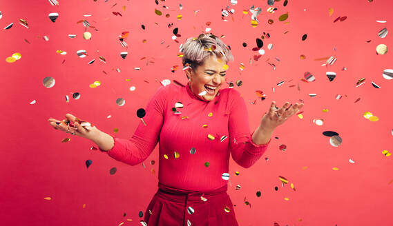 Time to celebrate. Carefree young woman getting excited about winning while standing under falling confetti in a studio. Vibrant young woman celebrating life and having fun against a red background. - JLPSF06291