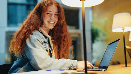 Porträt einer glücklichen jungen Studentin, die spät in der Nacht an ihrem Laptop lernt. Junge Frau, die einen Laptop am Bibliothekstisch benutzt und in die Kamera lächelt. - JLPSF06318
