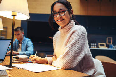 Confident girl preparing for her exams in library. Woman student sitting in library studying and smiling at camera. - JLPSF06321