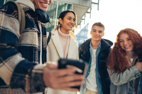 Gruppe von Menschen, die auf ein Smartphone in der Hand eines Freundes schauen. College-Studenten, die etwas Interessantes auf einem Mobiltelefon beobachten. - JLPSF06343