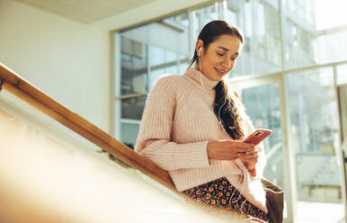 Studentin, die ihr Handy benutzt, während sie auf einer Treppe in der Universität steht. Junge Frau, die eine SMS auf einem Mobiltelefon auf dem College-Campus schreibt. - JLPSF06362