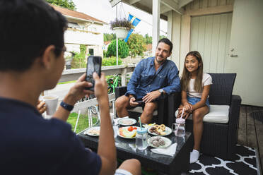 Boy photographing father and sister through smart phone sitting on chair at porch - DMMF00123