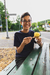 Glücklicher Junge, der auf einer Bank sitzend ein Eis genießt - DMMF00145