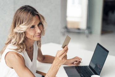 Businesswoman looking at credit card by laptop at office - JOSEF14143