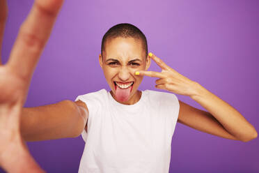 Portrait of expressive woman posing on purple background. Shaved head woman sticking out her tongue and showing peace hand sign. - JLPSF06536