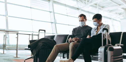 Tourists in face masks in airport wait for flight delayed or cancelled due to covid-19 lockdown. Travelers check airplane information online with smartphone in boarding lounge hall. - JLPSF06566