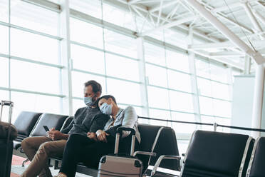 Man and woman with face masks sitting at airport waiting area. Couple at airport during Corona virus lockdown. - JLPSF06569