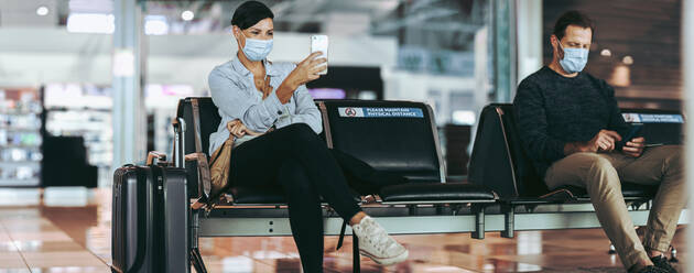 Man and woman traveler sitting apart maintaining social distance at airport. Male and female tourist with facemasks waiting at airport and using cell phone. - JLPSF06575