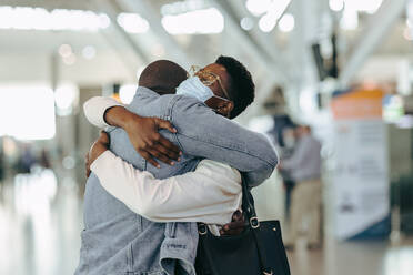 African traveler couple during pandemic meeting at airport. Man at airport terminal embracing woman with face mask before departure. - JLPSF06580
