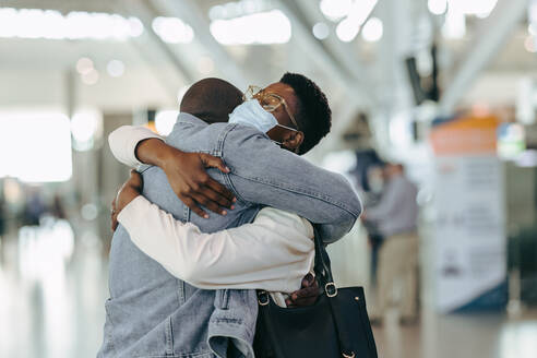 Afrikanisches Reisendenpaar während einer Pandemiebesprechung am Flughafen. Mann am Flughafenterminal umarmt Frau mit Gesichtsmaske vor dem Abflug. - JLPSF06580