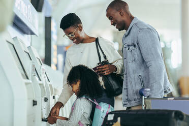 Afrikanische Familie, die ihre Bordkarte aus dem Check-in-Automaten am Flughafen ausdruckt. Dreiköpfige Familie auf dem Weg in den Urlaub am Flughafen. - JLPSF06581