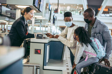 Reisende am Check-in-Schalter mit Mitarbeitern der Fluggesellschaft während der Pandemie. Afrikanische Familie beim Einchecken am Flughafen, Übergabe des Reisepasses an den Flugbegleiter. - JLPSF06583