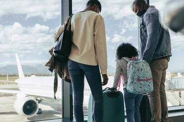 Rückansicht einer afrikanischen Familie, die mit ihrem Gepäck am Fenster des Flughafenterminals steht. Eine Touristenfamilie wartet mit ihrem Gepäck am Flughafen. - JLPSF06585