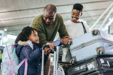 Touristisches Paar, das seine Tochter ansieht, während es mit seinem Gepäck am Flughafen steht. Afrikanisches Kind, das sich mit seinen Eltern im Flughafenterminal unterhält. - JLPSF06589