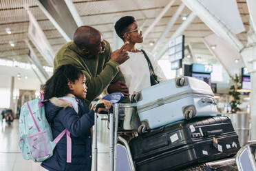 Ein Mann zeigt seiner Tochter und seiner Frau etwas, während er auf einem internationalen Flughafen steht. Eine dreiköpfige Familie wartet auf ihren Flug und schaut auf die Zeittafel. - JLPSF06590