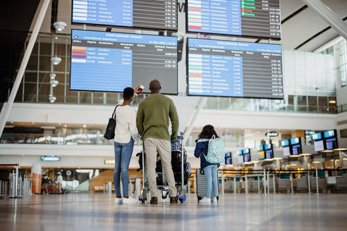 Rückansicht einer afrikanischen Familie mit Gepäck, die sich den Flugplan im Flughafenterminal ansieht. Eltern mit Tochter warten am Flughafen und überprüfen den Flugplan. - JLPSF06591