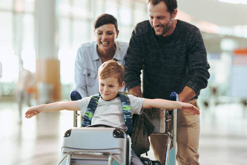 Boy sitting on luggage trolley while parents pushing it at airport. Family having fun at airport. - JLPSF06610