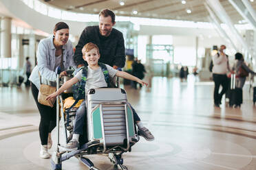 Eltern, die den Gepäckwagen mit ihrem Sohn am Flughafen bewegen. Ein Paar, das den Wagen mit seinem Sohn am Flughafen schiebt. - JLPSF06611