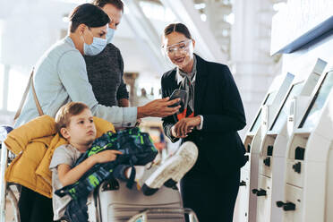 Ein Ehepaar zeigt dem Flugbegleiter am Flughafen sein Handy, während sein Sohn auf einem Gepäckwagen sitzt. Ein Flugbegleiter hilft einer Familie beim Self-Service-Check-in am Flughafen in Pandemie. - JLPSF06618