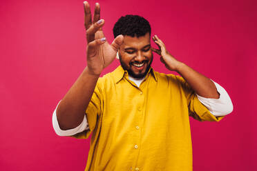 Streaming trendy music in the studio. Happy young man dancing cheerfully while listening to music using wireless earphones. Smiling young man standing alone against a pink background. - JLPSF06675