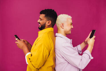 Carefree gay couple using their mobile phones in a studio. Happy young couple browsing the internet while standing back to back against a purple background. Cute gay couple enjoying being online. - JLPSF06689