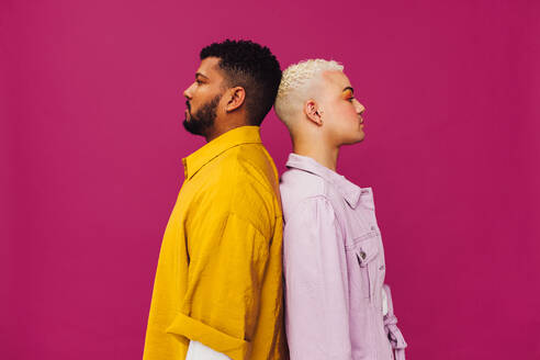 Confident young gay couple standing back to back in a studio. Sideview of a beautiful gay couple with piercings standing together against a purple background. - JLPSF06691