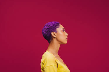 Seitenansicht einer modischen jungen Frau mit lila Haaren in einem Studio. Stilvolle junge Frau, die selbstbewusst vor einem rosa Hintergrund steht. - JLPSF06706