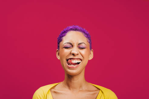 Aufgeregte junge Frau, die mit geschlossenen Augen ihre Zunge herausstreckt, während sie vor einem rosafarbenen Hintergrund steht. Modische junge Frau mit Make-up und lila Haaren in einem Studio. - JLPSF06708