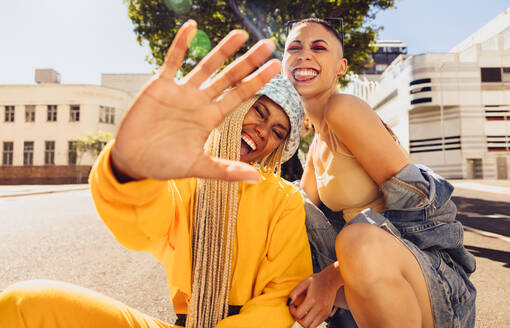 Friendship vibes only. Young trendy woman blocking the camera with her hand while sitting with her friend outdoors. Two best friends smiling cheerfully and having fun in the summer sun. - JLPSF06750