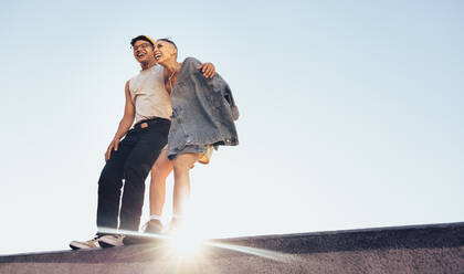 Zwei lebhafte Freunde lachen und haben Spaß im Freien. Zwei glückliche Freunde stehen auf einer Mauer mit dem Himmel im Hintergrund. Junger Mann und Frau fühlen sich energiegeladen und voller Leben. - JLPSF06832