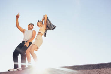 Zwei glückliche Jugendliche, die tanzen und sich im Freien amüsieren. Zwei energiegeladene junge Freunde, die auf einer Mauer stehen, mit dem Himmel im Hintergrund. - JLPSF06833