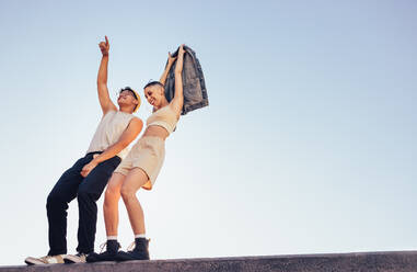 Hipster tanzen und haben Spaß im Freien. Zwei energiegeladene junge Leute stehen auf einer Mauer mit dem Himmel im Hintergrund. Zwei glückliche junge Freunde, die sich lebendig und voller Leben fühlen. - JLPSF06834