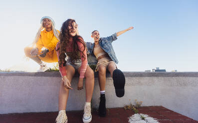Energiegeladene Freundinnen, die sich im Freien vergnügen. Drei glückliche Freundinnen, die lächelnd auf einer Mauer in der Stadt abhängen. Fröhliche weibliche Jugendliche, die zusammen Spaß haben. - JLPSF06851